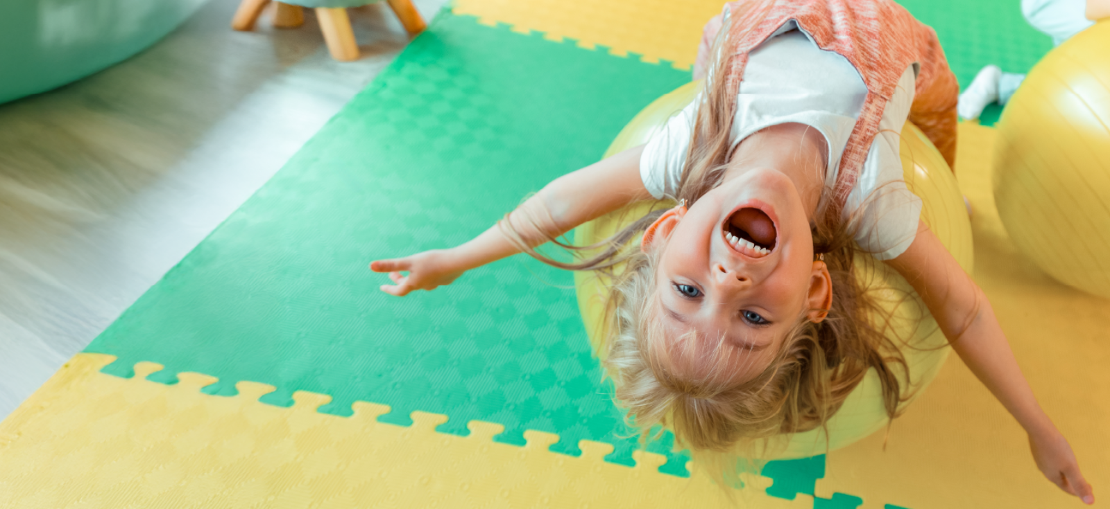 A child with a big happy smile is upside down on a yellow exercise ball