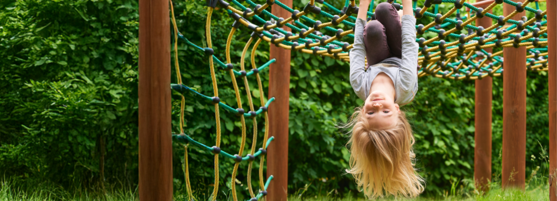 A young child is smiling while hanging upside down from a rope net climber outdoors