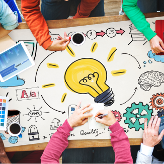 People collaborate around a table with a large paper displaying a lightbulb, gears, and charts, symbolizing brainstorming and innovation